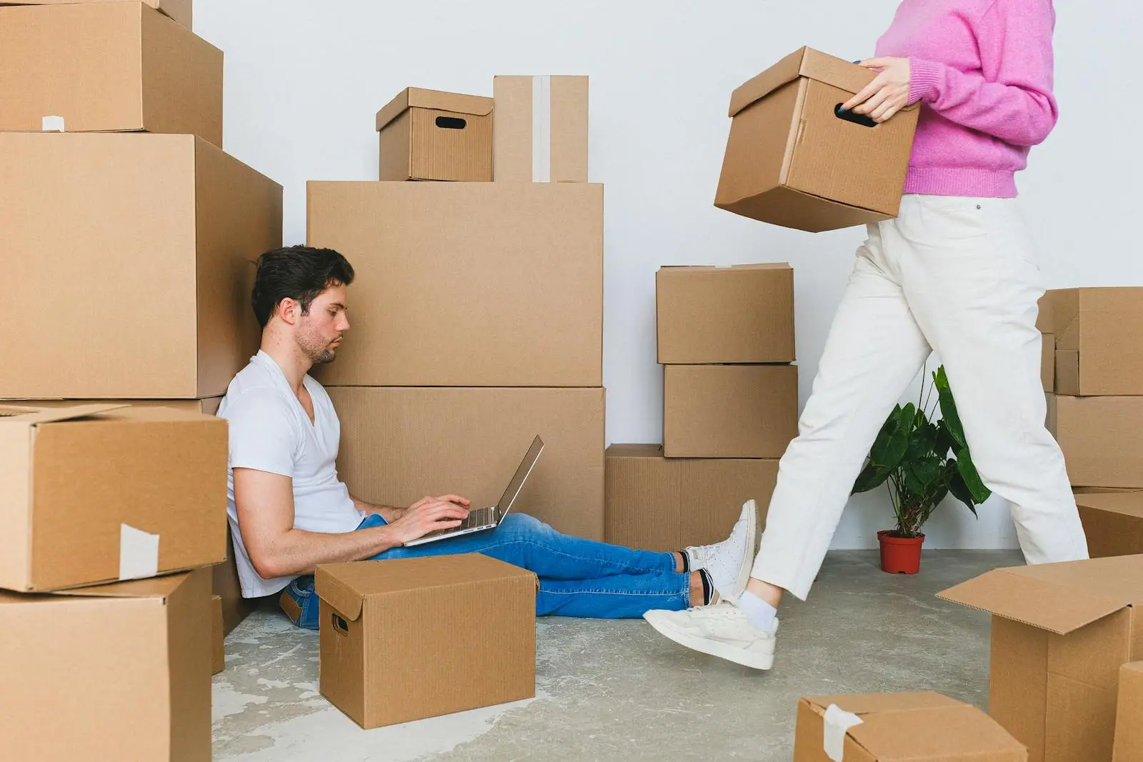 Crop woman arranging carton boxes during relocation with boyfriend using laptop on floor